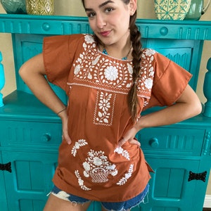 May include: A rust-colored embroidered blouse with white floral designs. The blouse has a round neckline and short sleeves.