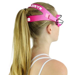 May include: A pink lacrosse goggle with the name "AURYN" printed in white on the headband. The number "8" is also visible. The goggle has a black cage and is designed to protect the eyes during play.