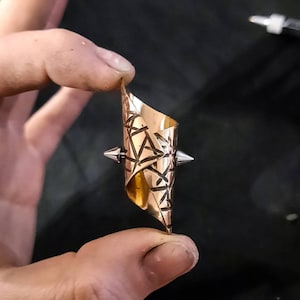 May include: A gold-colored ring with a geometric pattern and two silver spikes on the top and bottom. The ring is made of metal and has a unique design.
