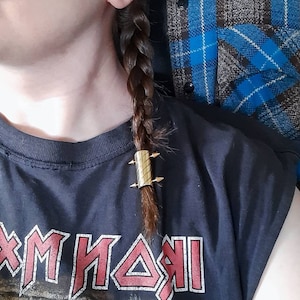 May include: A gold-colored hair tie with spikes, holding a braided ponytail. The hair tie is worn by a person wearing a black t-shirt with the text "Iron Maiden" printed on it.
