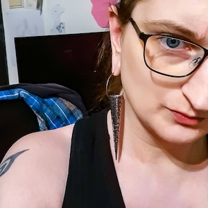 May include: A person wearing a black tank top and glasses with a long, silver, pointed earring. The earring is shaped like a cone with a wire design.
