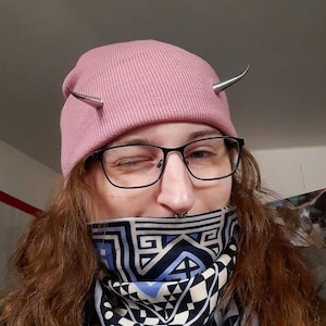 Könnte beinhalten: Eine Person trägt eine rosa Mütze mit silbernen Hörnern, eine schwarze Brille und eine schwarz-weiße Muster-Gesichtsmaske. Die Maske hat ein geometrisches Design mit blauen Akzenten.