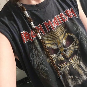 Peut inclure: Une personne portant un t-shirt noir avec les mots "Iron Maiden" imprimés dessus. La personne a une tresse dans les cheveux avec un clip à cheveux argenté avec des pointes.