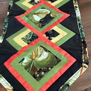 May include: Quilted table runner with a repeating diamond pattern. The design uses black, green, yellow, and red fabrics. The central diamonds display a large-leaf floral print, creating a vibrant, decorative accent for any table setting.