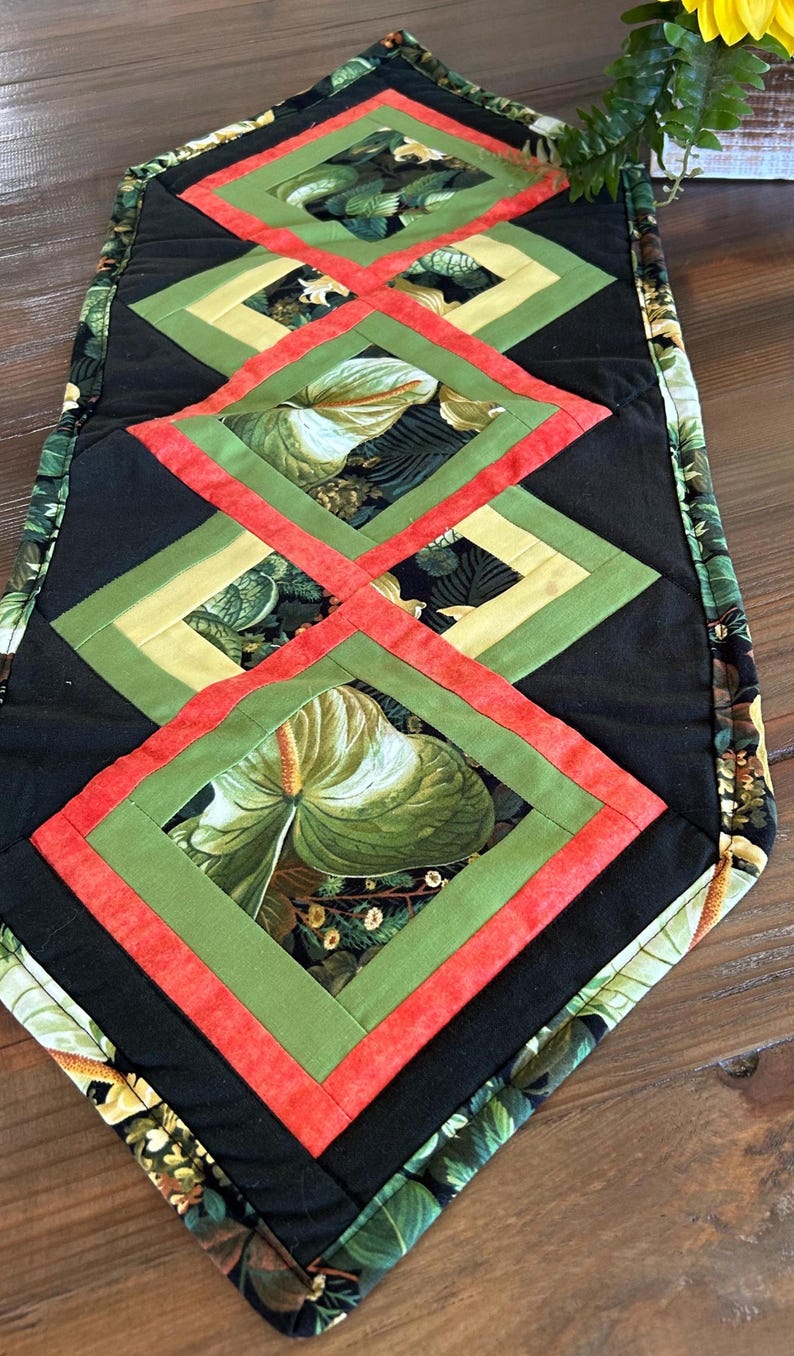 May include: Quilted table runner with a repeating diamond pattern. The design uses black, green, yellow, and red fabrics. The central diamonds display a large-leaf floral print, creating a vibrant, decorative accent for any table setting.