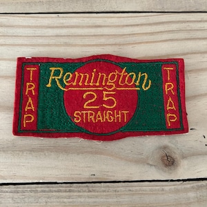 May include: A rectangular patch with the words "Remington 25 STRAIGHT" embroidered in yellow on a red and green background. The word "TRAP" is embroidered vertically on both sides.
