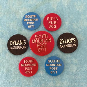 May include: A collection of seven circular tokens with printed text. The tokens are in various colors, including red, black, and blue. The text includes "SOUTH MOUNTAIN POST 6771", "SID'S PUB 303", and "DYLAN'S EAST BERLIN, PA".