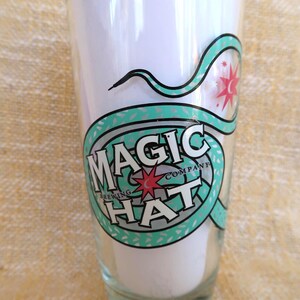 May include: A clear glass with the words "MAGIC HAT BREWING COMPANY" in a circular design. The design is in teal, black, and white, with a red starburst. The glass is filled with a white substance.