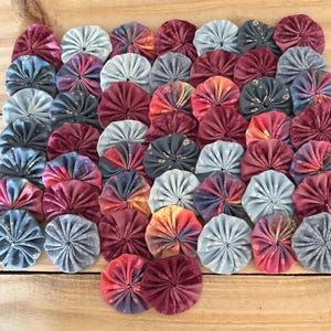 May include: A collection of colorful, circular fabric pieces arranged on a wooden surface. The pieces are in shades of blue, pink, red, and gold, with a pleated, flower-like design. The arrangement is dense, with the pieces closely packed together.