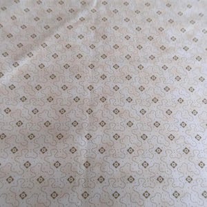May include: Close-up of a cream-colored fabric with a repeating pattern of small brown diamonds and dotted lines. The fabric has a slightly wrinkled texture, suggesting it may be cotton or linen. The design is simple and elegant.