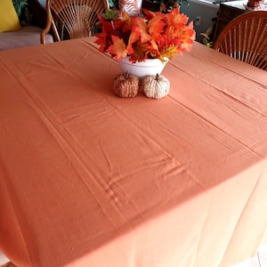 May include: A large, rectangular orange tablecloth covers a table. A white bowl filled with orange and red autumn leaves sits in the center, with two decorative pumpkins. The scene evokes a fall harvest theme.
