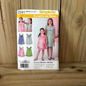 May include: A Simplicity sewing pattern envelope labeled 2241, size K5 7, 8, 10, 12, 14. The front features illustrations and photographs of sleeveless dresses with ruffles and pockets. Text includes Learn to Sew and Learn Basics Skills.