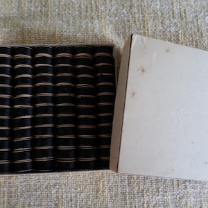 May include: A box of black hair rollers with tan bands. The rollers are neatly arranged inside a rectangular cardboard box with a detached lid. The box and rollers are set against a textured, light-colored background.
