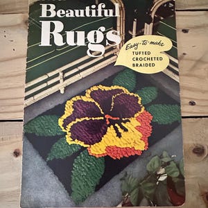 May include: A vintage instructional booklet titled Beautiful Rugs, Star Book No. 73, featuring a cover illustration of a pansy-shaped rug with purple, yellow, and orange petals on a black background, with text reading Easy-to-make TUFTED CROCHETED BRAIDED.