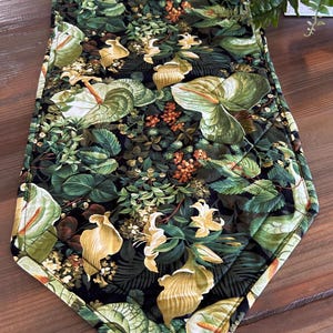 May include: A quilted table runner with a botanical print featuring green leaves, yellow calla lilies, and small orange flowers on a black background. The runner has a diamond-stitched pattern and a dark border.