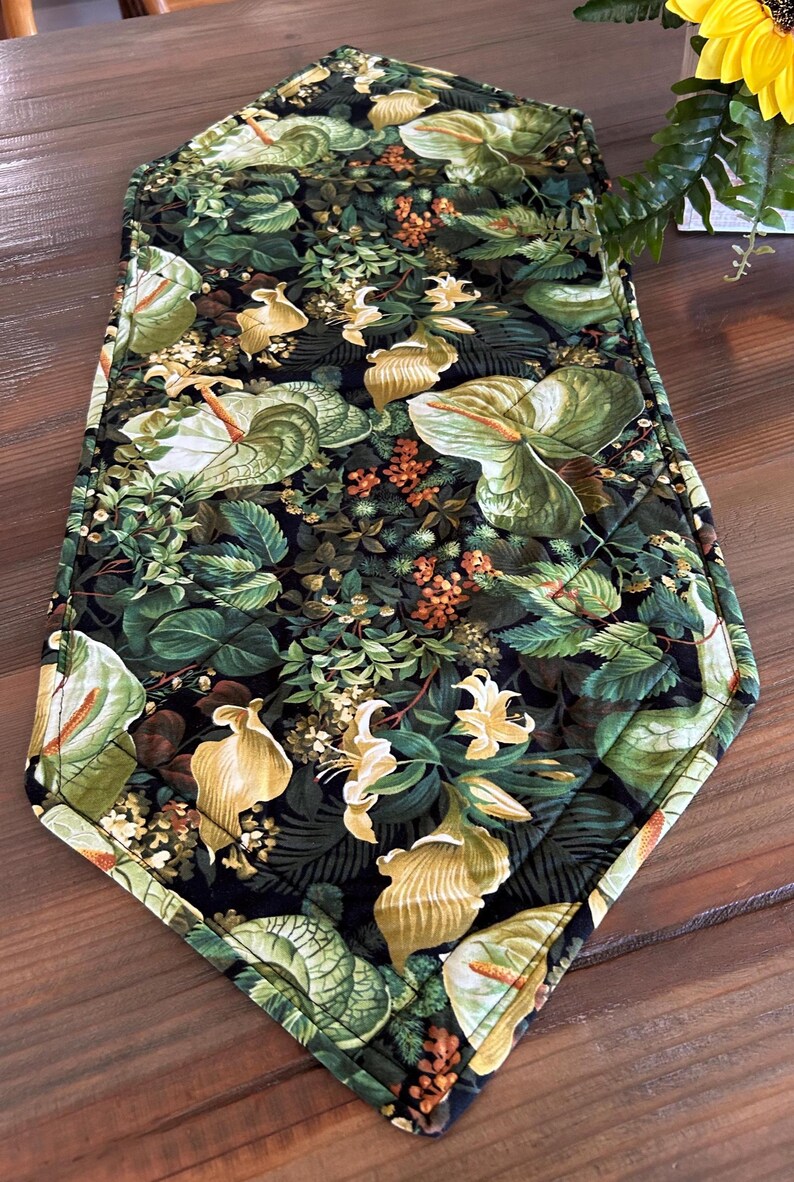 May include: A quilted table runner with a botanical print featuring green leaves, yellow calla lilies, and small orange flowers on a black background. The runner has a diamond-stitched pattern and a dark border.