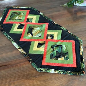 May include: A quilted table runner with a black background and square floral designs in red, green, and yellow. The runner's border features a green and black floral pattern. The runner is placed on a wooden surface.