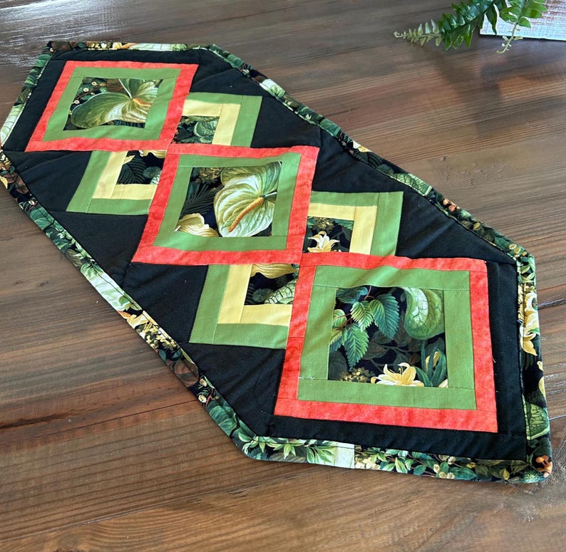 May include: A quilted table runner with a black background and square floral designs in red, green, and yellow. The runner's border features a green and black floral pattern. The runner is placed on a wooden surface.
