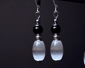 May include: A pair of dangle earrings with a black bead, silver accents, and a white oval-shaped stone. The earrings hang from silver hooks against a dark background. The jewelry is elegant and simple.