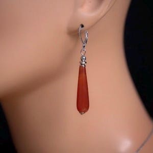 May include: A drop earring featuring a smooth, elongated, teardrop-shaped orange carnelian stone suspended from a silver-toned metal leverback hook. The stone is accented by a small, round, ridged silver-toned metal bead at the top of the drop.