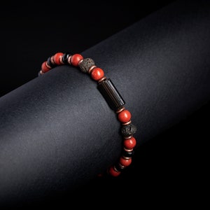 Peut inclure: Un bracelet de perles rouges, noires et bronze. Le bracelet comprend des perles rondes rouges, des perles cylindriques noires et des perles texturées de couleur bronze. Le bracelet est présenté sur un fond noir.