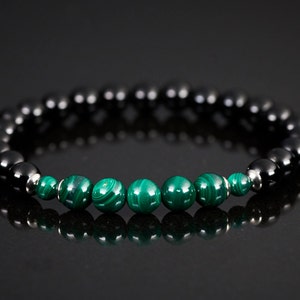 May include: A stretch bracelet with black and green malachite beads. The bracelet is displayed on a reflective black surface. The green malachite beads have a distinctive swirl pattern. The bracelet is a stylish accessory.