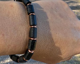 May include: Black gemstone bracelet with copper-colored metal accents. The bracelet features rectangular, faceted black stones separated by small copper-colored rings. The bracelet is worn on a wrist.