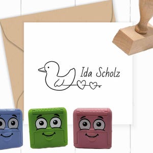 Op de afbeelding: Een briefpapier set met een witte kaart met een eendenillustratie en de naam "Ida Scholz", een bruine envelop, een houten stempel en drie gekleurde zelfinktende stempels in blauw, groen en roze, elk met een lachend gezicht.