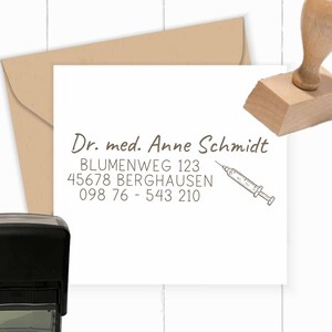 May include: A brown self-inking rubber stamp with the text "Dr. med. Anne Schmidt" and the address "Blumenweg 123, 45678 Berghausen, 098 76 - 543 210" and a small illustration of a syringe.