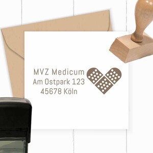 May include: A brown rubber stamp with a heart-shaped design featuring two plasters. The text "MVZ Medicum Am Ostpark 123 45678 Köln" is printed below the heart.