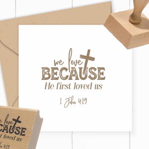 May include: A wooden rubber stamp featuring the text 'we love BECAUSE He first loved us 1 John 4:19' with a cross symbol. The stamp is shown alongside a square white card displaying the same printed design in brown ink and a tan envelope.