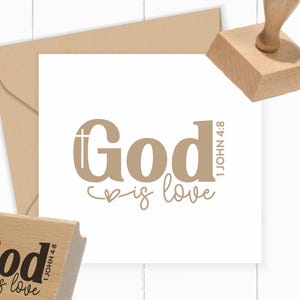 Könnte beinhalten: Ein Holzstempel mit dem Text God is love und 1 John 4:8, wobei ein Kreuz in den Buchstaben G integriert ist. Der Stempel wird zusammen mit einem beigen Umschlag und einer weißen quadratischen Karte gezeigt, die das Motiv in hellbrauner Tinte trägt.