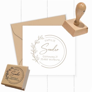 May include: A wooden self-inking stamp with a brown ink pad. The stamp features a circular design with a wreath of leaves and the text "Familie Sander" in a cursive font. The address "Gotheweg 21, 35468 Wulfburg" is printed below the name.