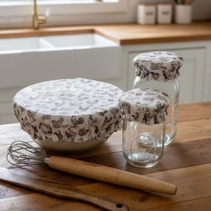 Op de afbeelding: Een keuken met een witte kom en twee glazen potten, bedekt met stoffen deksels met kippenmotieven. Een garde en een houten spatel liggen op een houten oppervlak. De achtergrond toont een gootsteen, kasten en een raam.