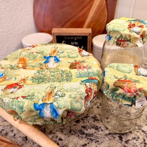May include: Reusable food covers with a Peter Rabbit design. The covers are yellow with illustrations of Peter Rabbit and other characters. The covers are on glass jars and a wooden rolling pin.