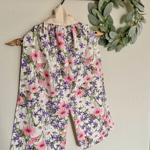 May include: A floral kitchen towel with a white background, featuring pink and purple flowers. The towel is hanging on a wooden rack. A decorative wreath is visible in the background.