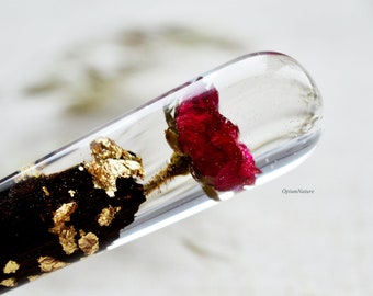 May include: A clear glass tube filled with resin, containing a single red rose and gold flakes. The rose is preserved in the resin, and the gold flakes add a touch of sparkle.