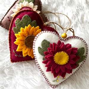 Op de afbeelding: Drie vilten hartvormige ornamenten met geborduurde bloemen. De harten zijn wit, rood en bruin. De bloemen zijn geel en rood.