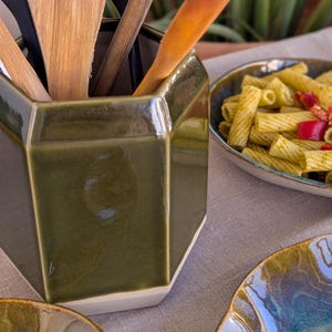 Può includere: Un portautensili esagonale in ceramica verde oliva con base bianca, contenente diversi utensili da cucina in legno tra cui una spatola forata e cucchiai, posizionato accanto a un piatto di pasta con peperoni rossi e una ciotola in ceramica blu.
