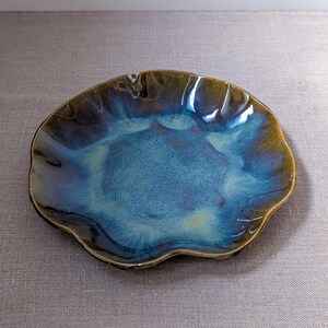 Op de afbeelding: Een handgemaakte keramische schaal met een unieke, organische vorm. De schaal heeft een glanzend, iriserend glazuur met tinten blauw, groen en bruin. De rand heeft een donkerdere kleur en de schaal heeft een diameter van ongeveer 20 cm.