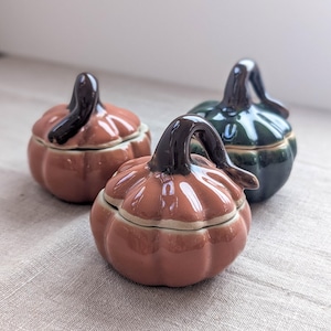 Pode incluir: Três recipientes de cerâmica em forma de abóbora com tampa e acabamento brilhante. Dois recipientes são de um laranja terracota suave e um é de um verde floresta profundo. Cada um apresenta uma alça de caule curva castanha escura.