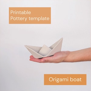 Op de afbeelding: Een lichtgrijze keramische sculptuur in de vorm van een gevouwen papieren bootje. De afbeelding bevat de tekst "Printable Pottery template" en "Origami boat" op oranje rechthoekige achtergronden. Het object heeft een gladde, matte afwerking en puntige uiteinden.