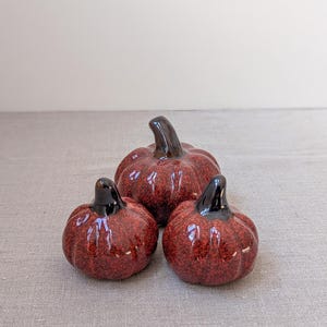 Op de afbeelding: Drie kleine keramische pompoenen met een glanzende, gespikkelde dieprode afwerking en gladde, donkerbruine stelen. De pompoenen hebben een ronde, geribbelde vorm met een reflecterend oppervlak en zijn in een groepje bij elkaar geplaatst.