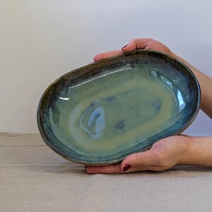May include: An oval ceramic dish featuring a glossy glaze in shades of sage green and deep blue. The interior surface displays a smooth, reflective finish with subtle color variations, while the raised edges are finished in a darker, earthy tone.