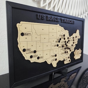 Pu&ograve; includere: Una mappa in legno degli Stati Uniti con buchi neri che rappresentano i circuiti di gara. La mappa &egrave; incorniciata in nero e ha il testo "US RACE TRACKS" in alto.