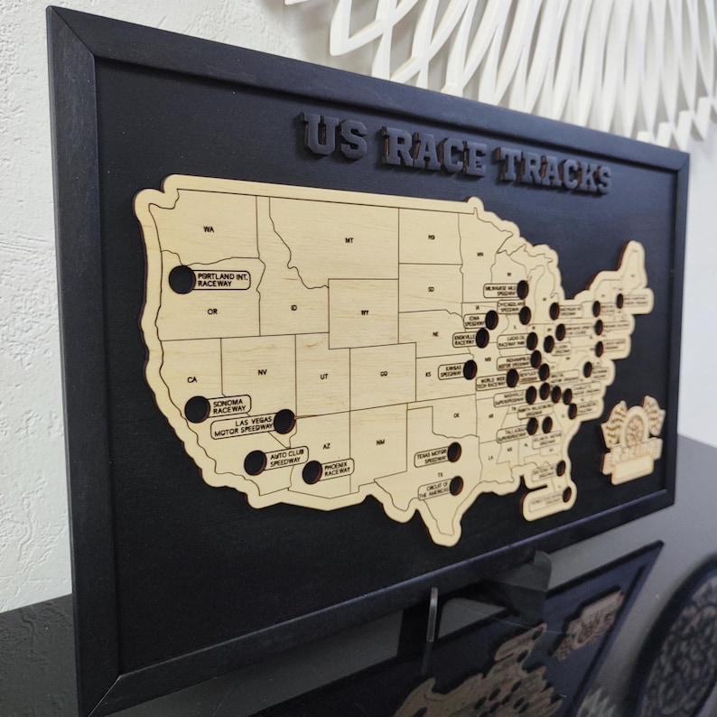 Pu&ograve; includere: Una mappa in legno degli Stati Uniti con buchi neri che rappresentano i circuiti di gara. La mappa &egrave; incorniciata in nero e ha il testo "US RACE TRACKS" in alto.