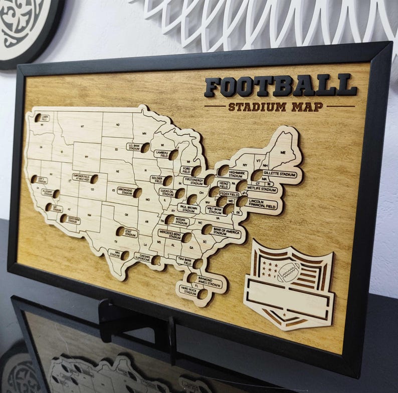 May include: A wooden map of the United States with the names of football stadiums in each state. The map is framed in black and has a football-shaped logo in the bottom right corner. The text "FOOTBALL STADIUM MAP" is at the top of the map.