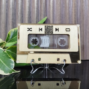 May include: A gold-colored wooden cassette tape with a clear plastic window. The cassette has a QR code, rewind, play, fast forward, and stop buttons. The cassette is on a clear plastic stand.