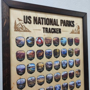 May include: A wooden US National Parks Tracker board in a dark brown frame. The light wood surface features the laser-cut text US NATIONAL PARKS TRACKER and rows of colorful shield-shaped stickers representing various national parks with names.