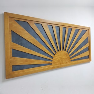 Puede incluir: Una pieza de arte mural rectangular con un marco de madera marrón claro y un diseño de rayos de sol. El diseño consiste en vigas de madera radiantes en un acabado marrón claro sobre un fondo azul oscuro, creando un patrón de alto contraste.